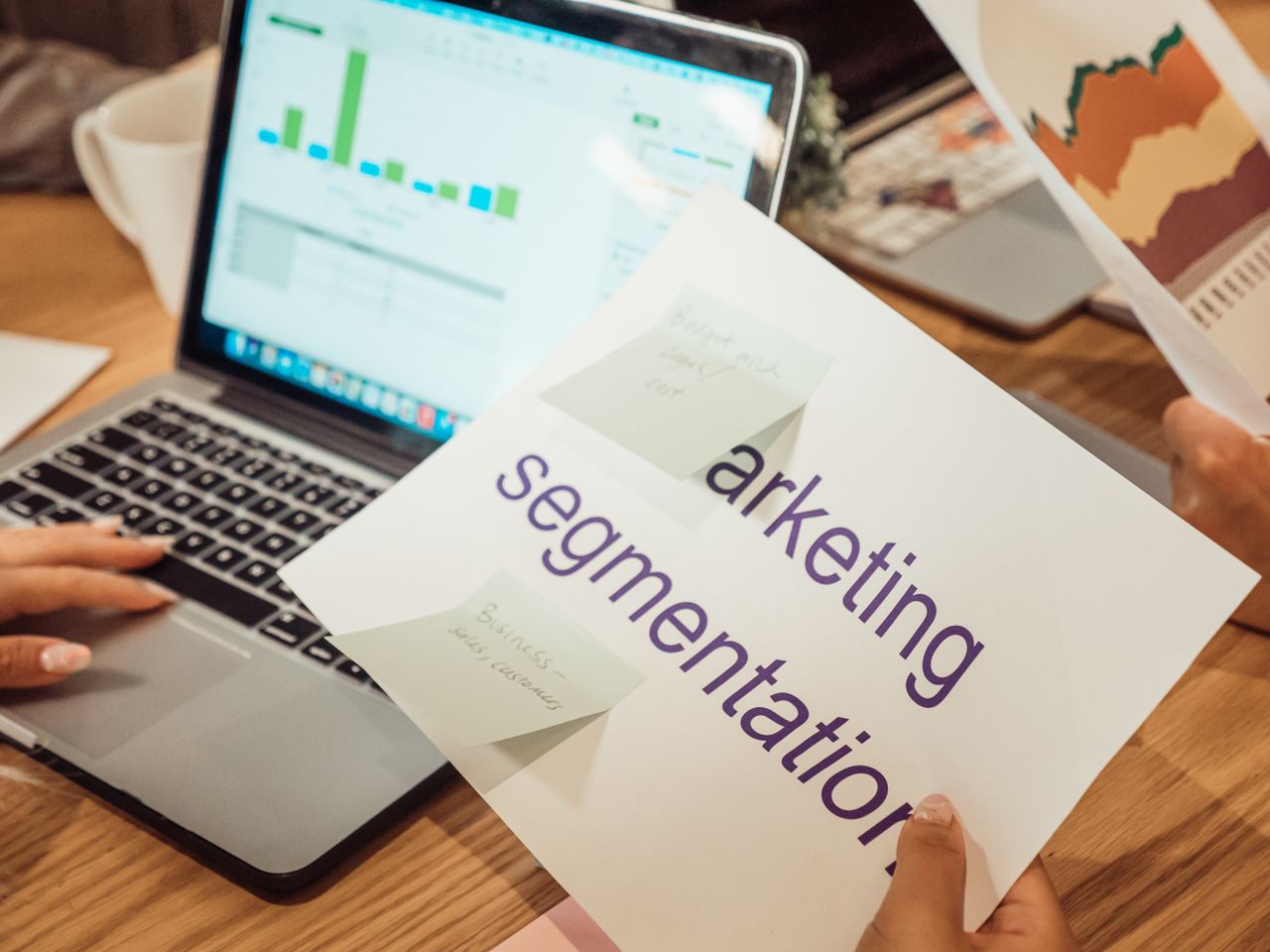 A contemporary office setting with hands reviewing marketing data on a laptop and printed documents.